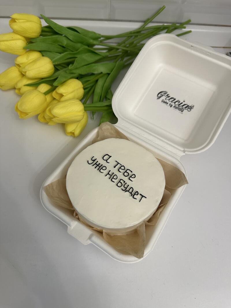A small white cake with the inscription "а тебе уже не будет" in black lettering is nestled in an open white container, accompanied by a bouquet of yellow tulips.