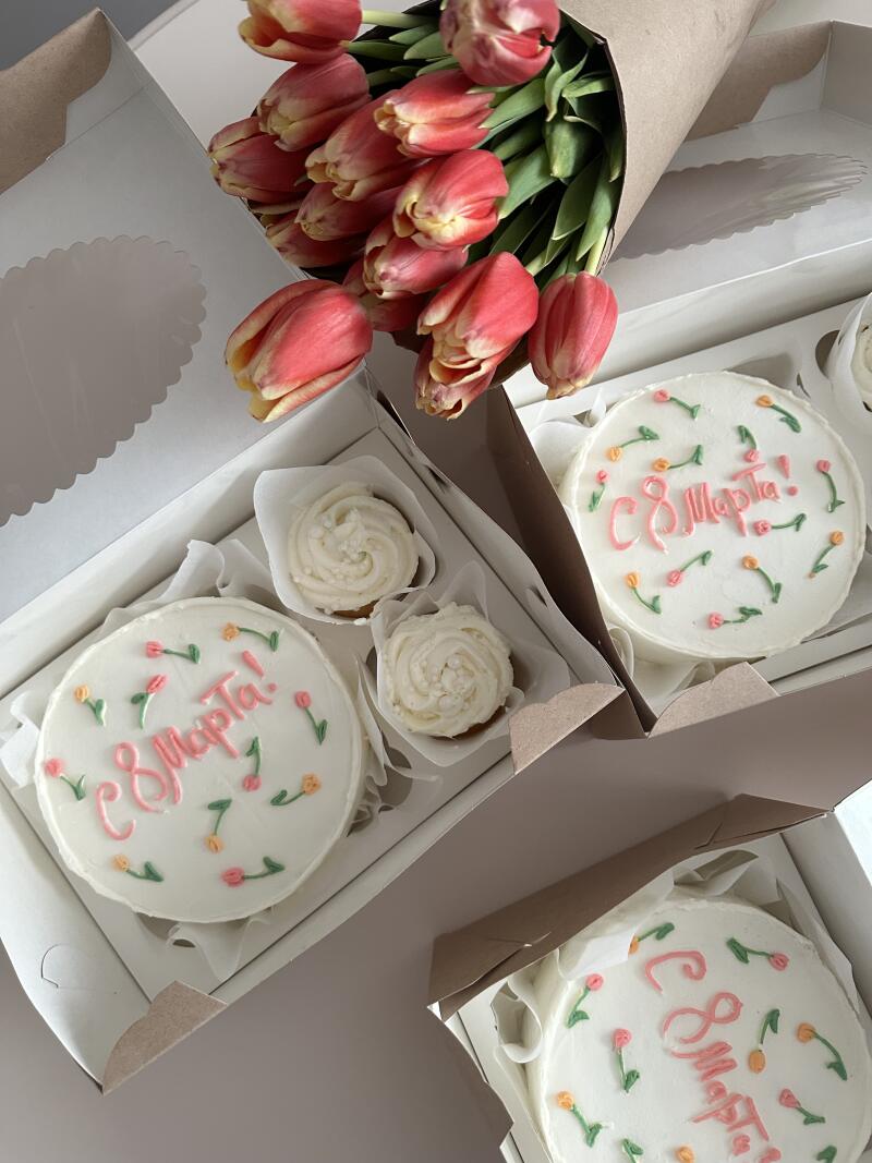 The image shows a bouquet of tulips and several boxes, each containing a small cake decorated with the inscription "С 8 Марта!" and two cupcakes.