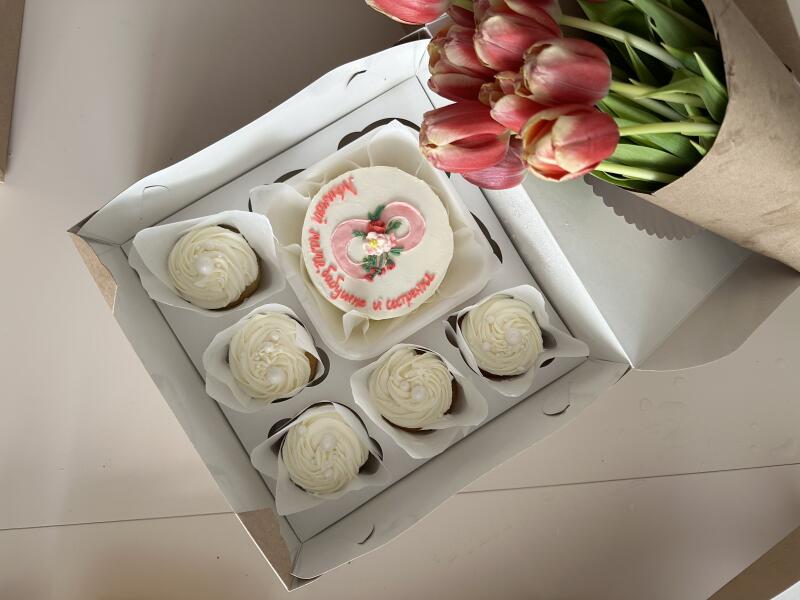 A box of cupcakes with white frosting and a small cake decorated with flowers and the inscription "To my beloved grandmother and sister" sits next to a bouquet of pink tulips wrapped in brown paper.