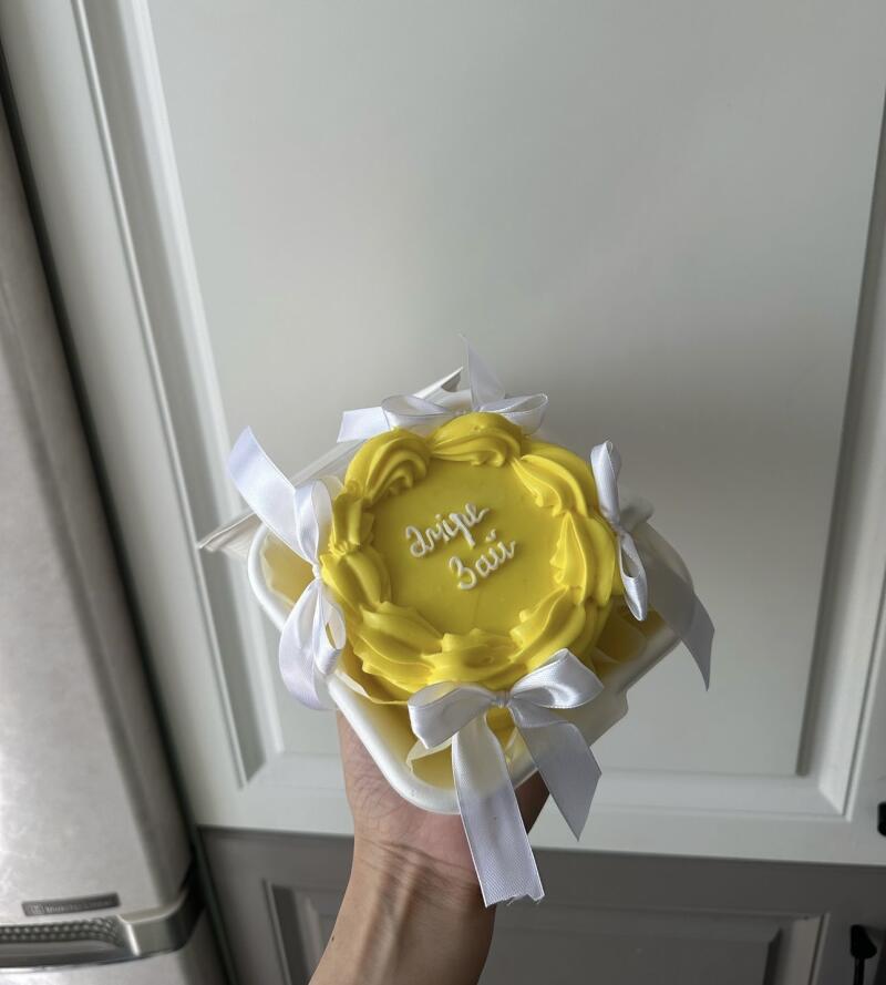 A hand holds a small yellow cake decorated with white ribbons and the inscription "Аміре 3 жай".