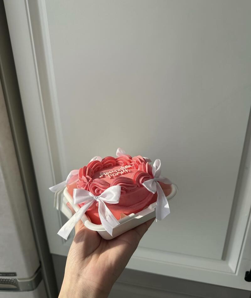 A hand holds a small, square pink cake decorated with pink frosting swirls and white ribbons, presented in a white container; text on the cake reads "С Днем Рождения!"