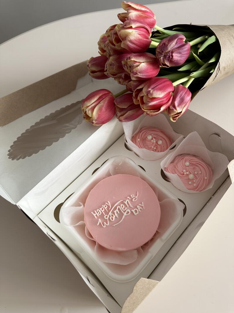 A box containing a cake with the inscription "Happy Women's DAY" and two cupcakes is next to a bouquet of pink tulips wrapped in paper.