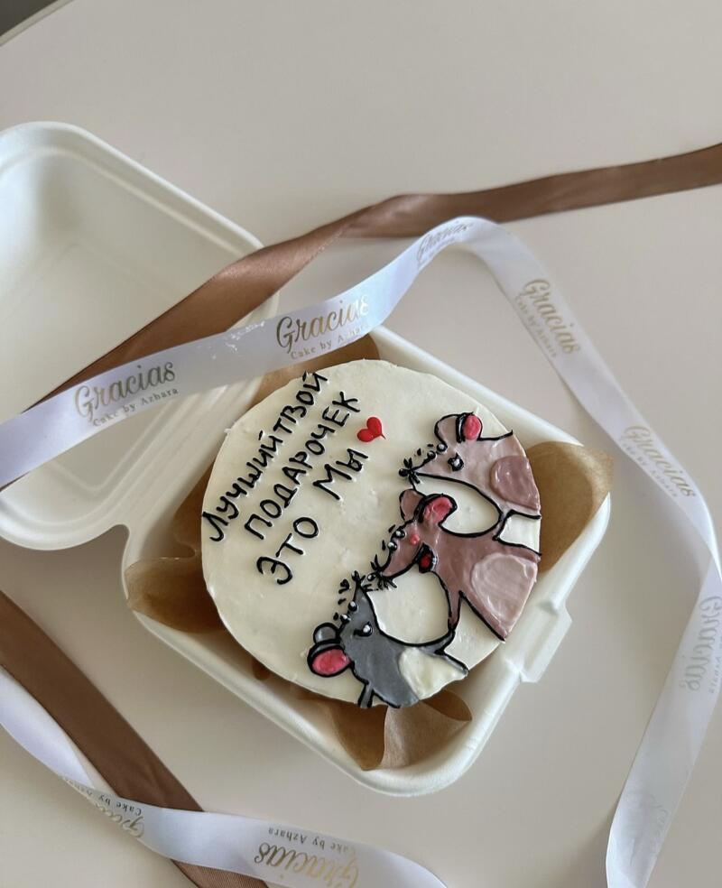 A small round cake decorated with three cartoon mice and the inscription "The best gift is us" in Russian sits in a white clamshell container, surrounded by white ribbon with the text "Gracias Cake by Azhara" and a brown ribbon.
