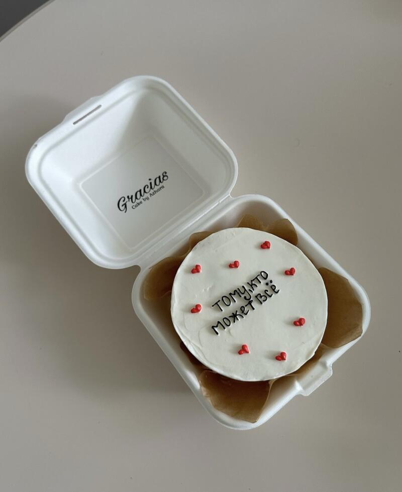 A small cake with white frosting, red heart decorations, and the Russian text "Тому, кто может всё" inside a white take-out container with the logo "Gracias Cake by Azhara".