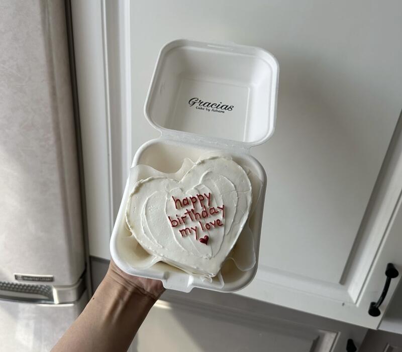 A hand holds a heart-shaped cake with white frosting inside an open white container; the cake has "happy birthday my love" written in red icing and a small red heart.