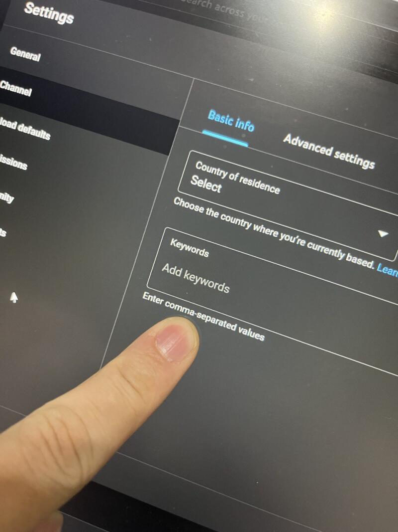 The image shows a screen displaying settings, specifically the 'Basic info' section for a channel. The options visible include 'Country of residence,' 'Keywords' with a prompt to 'Add keywords,' and the instruction 'Enter comma-separated values,' indicated by a finger pointing towards it.