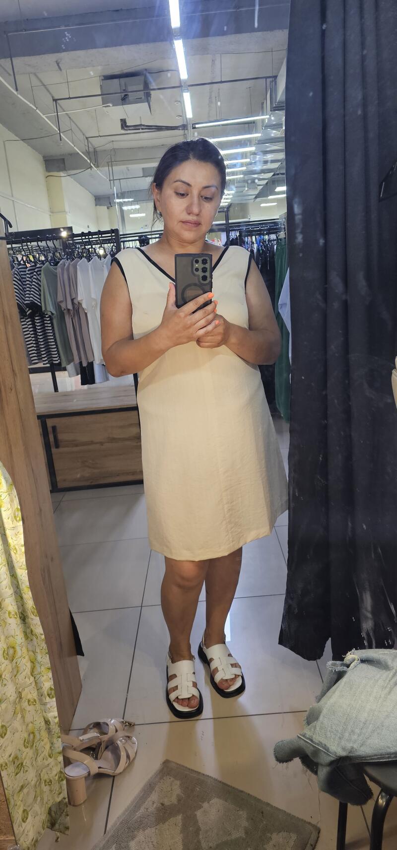 A woman takes a mirror selfie wearing a cream-colored dress with black trim and white platform sandals.