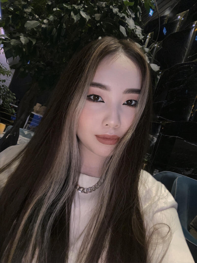 A young woman with long, highlighted hair and winged eyeliner wears a silver chain necklace and a light-colored shirt.