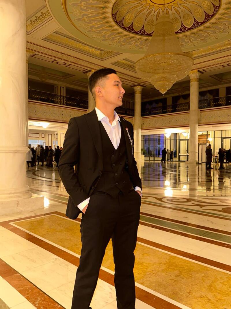 A man in a black suit stands in a large hall with a chandelier and marble floors.