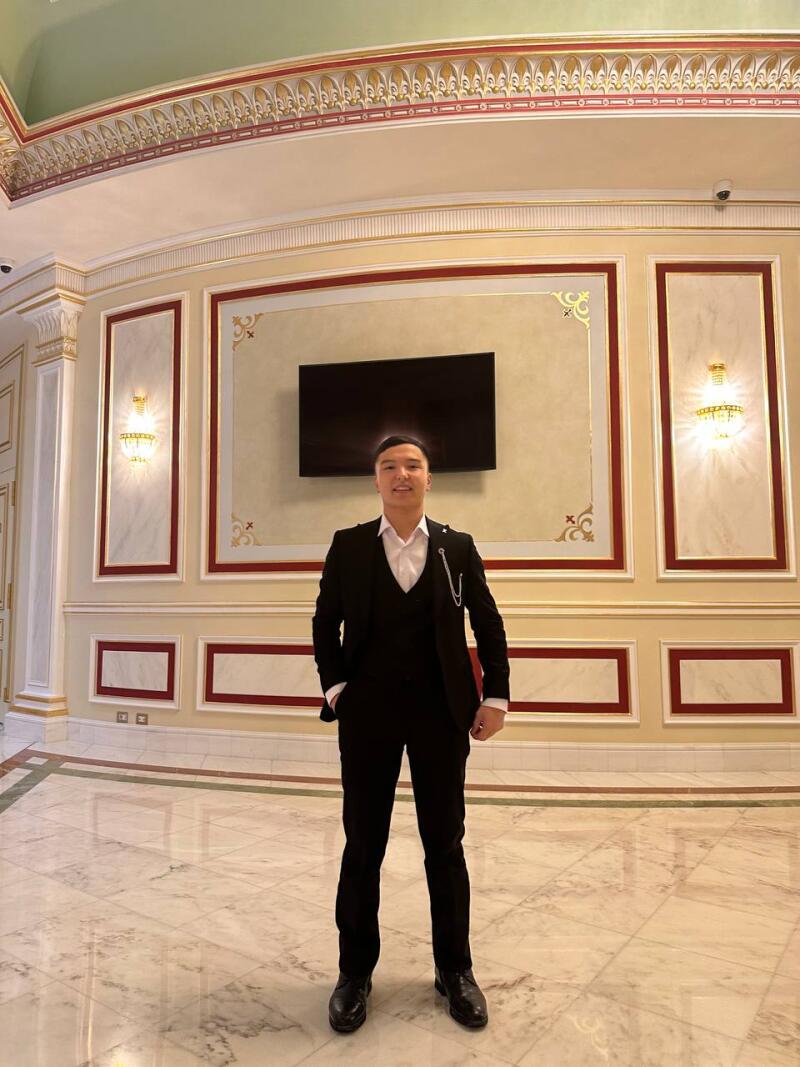 A man in a black suit stands in a room with ornate walls and marble floors.
