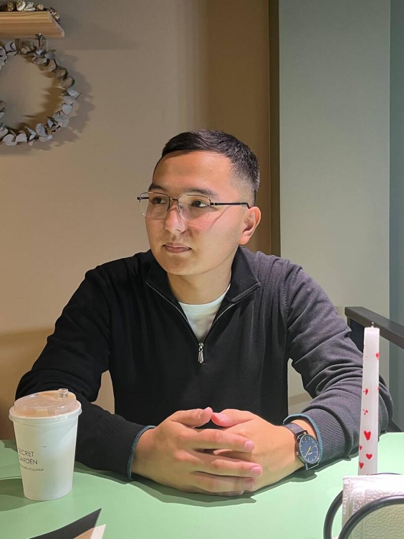 A man wearing glasses and a black sweater sits at a green table with a coffee cup and a candle decorated with red hearts.