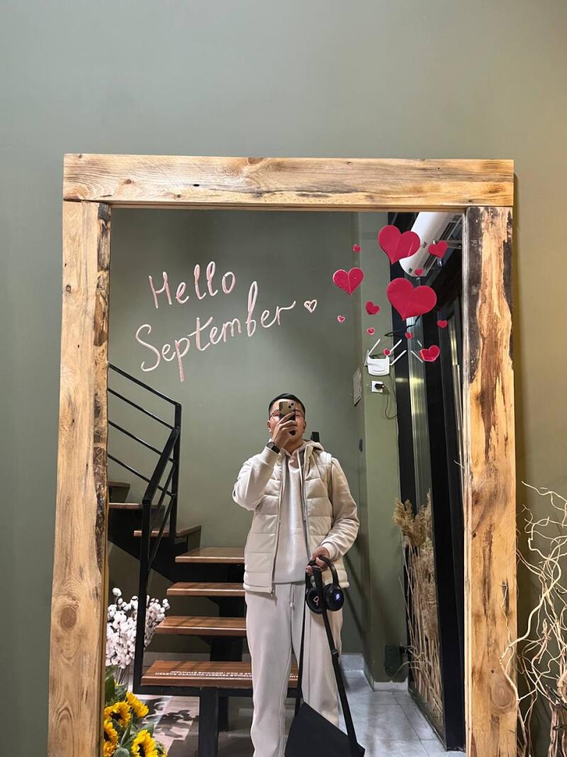 A person in a cream-colored outfit takes a mirror selfie in a wooden-framed mirror with red hearts and the text “Hello September” on it.