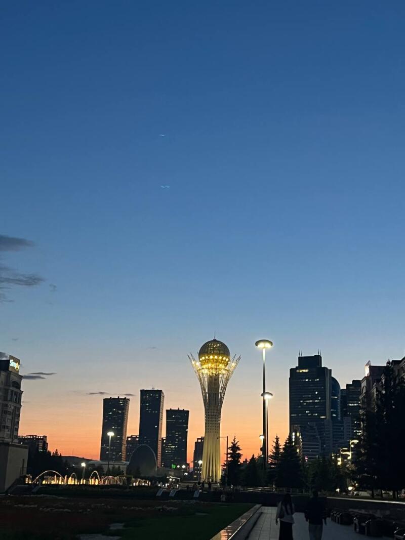 The image shows the Bayterek monument in Nur-Sultan at dusk, with skyscrapers in the background.