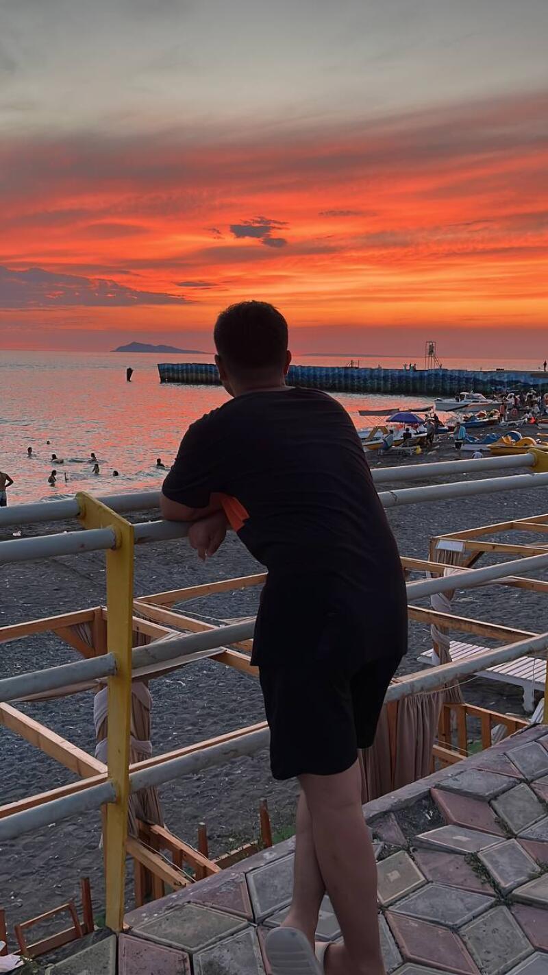 A person in black clothes is leaning on a railing, looking out at a beach at sunset with orange and red skies.