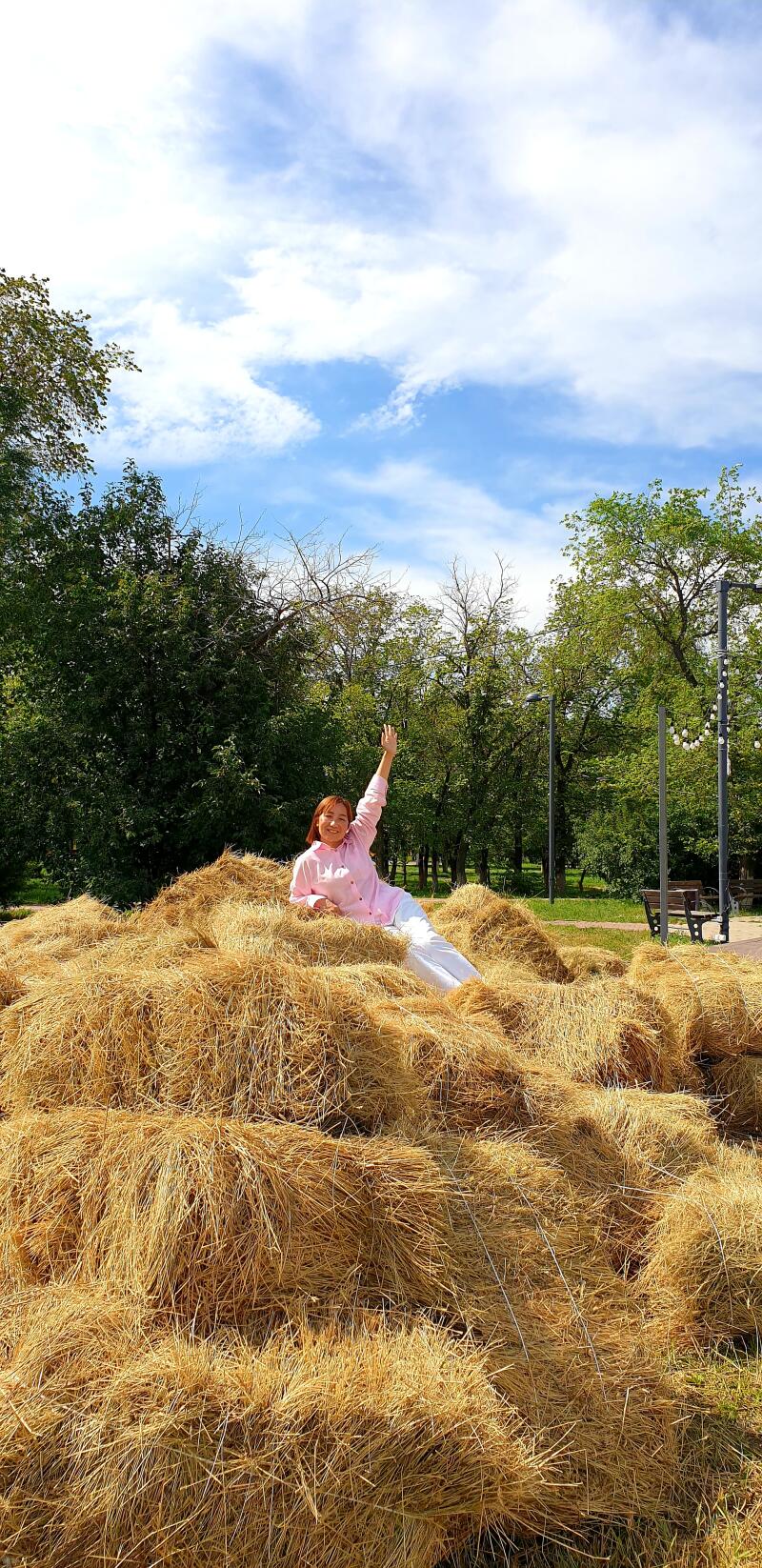 A woman in a pink shirt and white pants is lying on a stack of hay bales with her arm raised in the air, against a backdrop of trees and a partly cloudy sky.