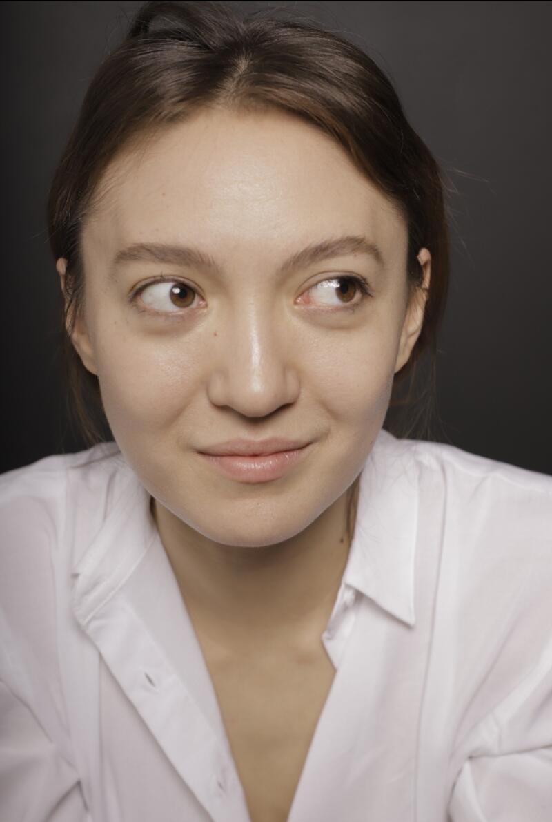 A woman with brown hair and eyes is wearing a white collared shirt, looking to the side with a slight smile against a dark background.