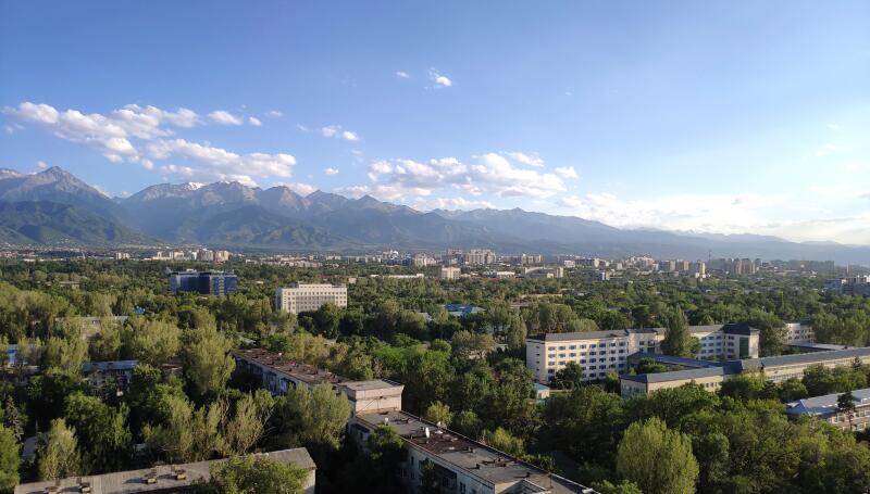 An aerial view shows a city with numerous buildings and trees, backed by a mountain range under a partly cloudy sky.