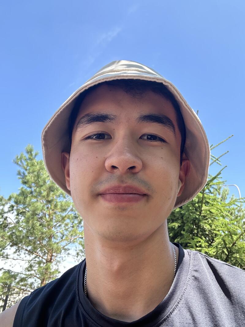 A selfie of a person wearing a beige bucket hat and a black tank top with a silver chain, with trees and a blue sky in the background.