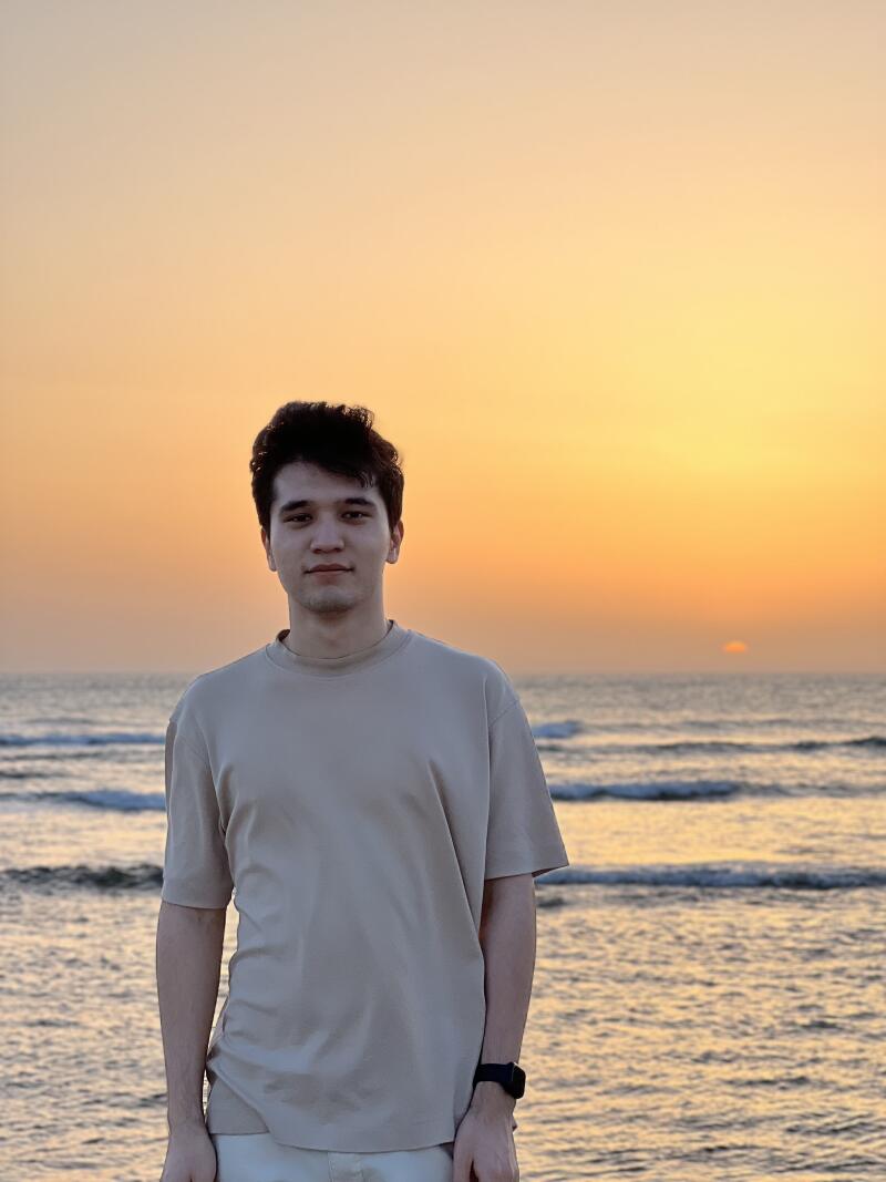 A man in a beige T-shirt stands in front of the ocean at sunset, wearing a black watch.