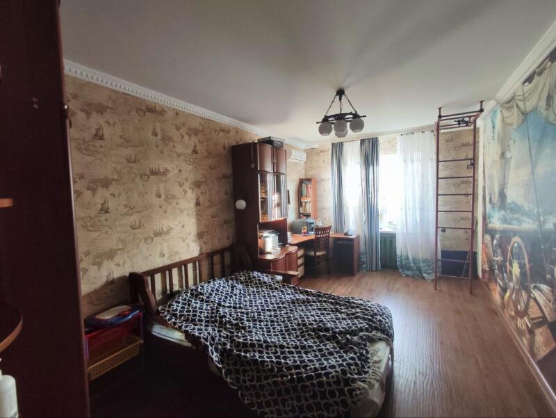 A bedroom features a bed with a patterned blanket, a desk with a chair, and a wall-mounted ladder.
