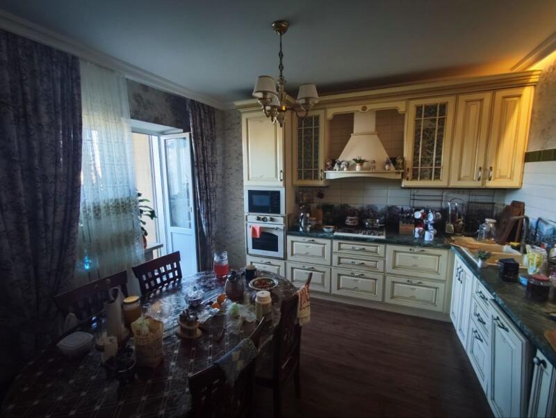The photo shows a kitchen with light-colored cabinets, a dark wood floor, and a dining table covered with food and various items.