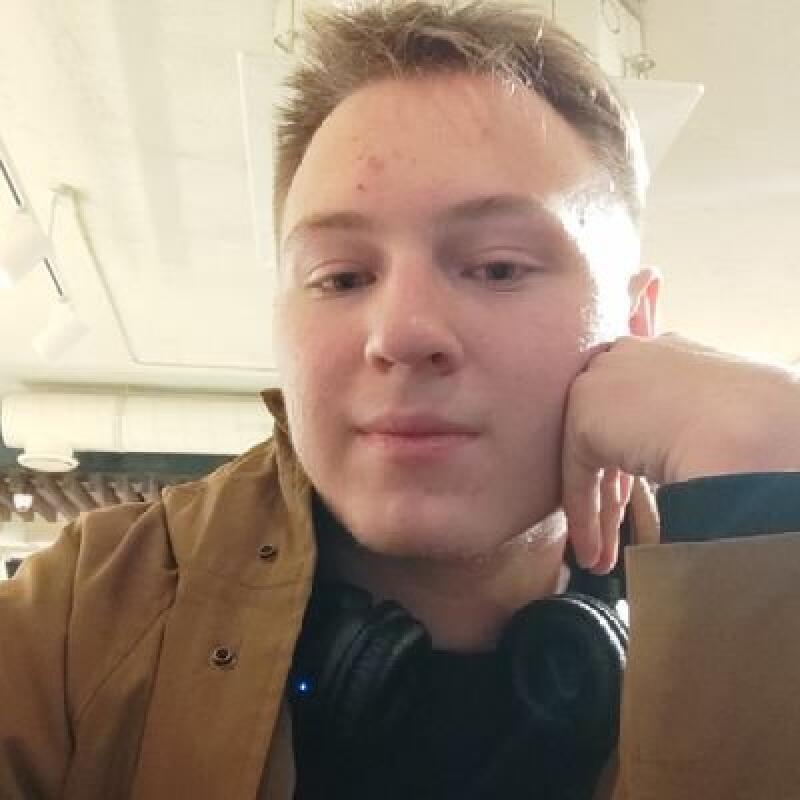 A man is posing with his hand on his cheek, wearing headphones around his neck and a brown jacket.