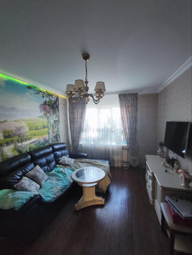 A living room features a black leather sofa with patterned cushions and a turquoise blanket, a round white table, and a TV on a light-colored cabinet, with a landscape mural and a chandelier.
