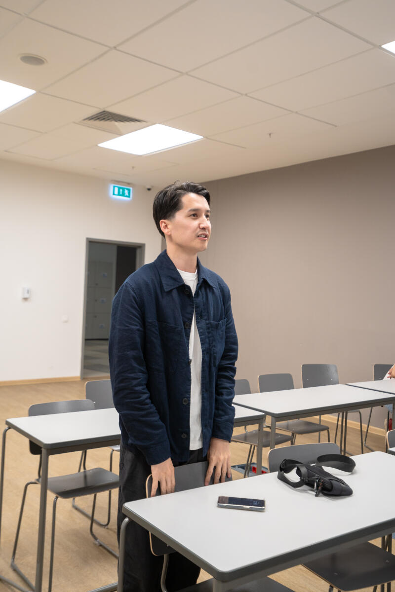 A man in a blue jacket and white t-shirt stands in a classroom, with a phone and a small black bag on the table in front of him.