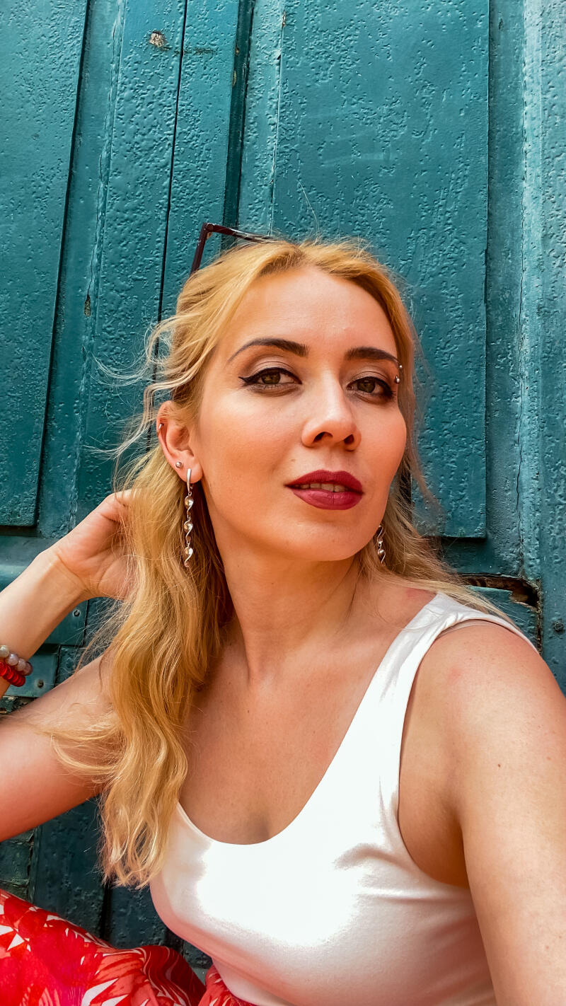 A woman with blonde hair, dark eyeliner, and dark red lipstick is wearing a white top and silver earrings against a teal textured background.