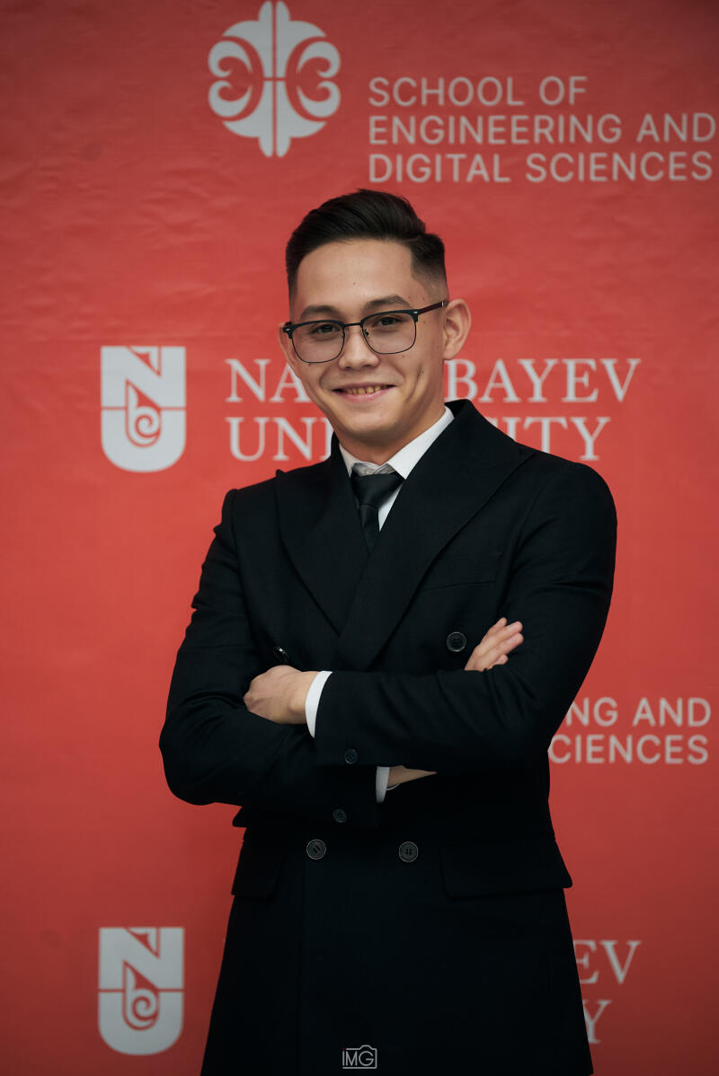 A smiling man wearing glasses and a black suit stands with his arms crossed in front of a red backdrop with the words "SCHOOL OF ENGINEERING AND DIGITAL SCIENCES" and "NAZARBAYEV UNIVERSITY" printed on it.