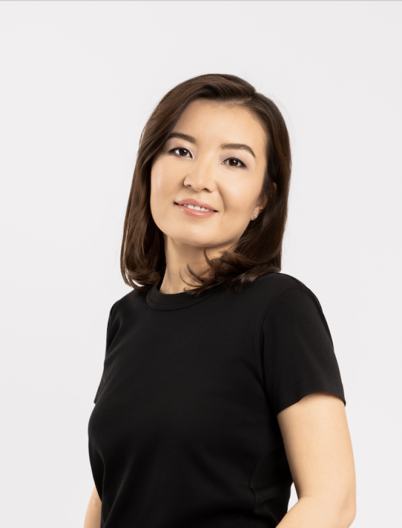 A woman with dark brown hair and a slight smile looks directly at the camera, wearing a black short-sleeved t-shirt against a plain light background.
