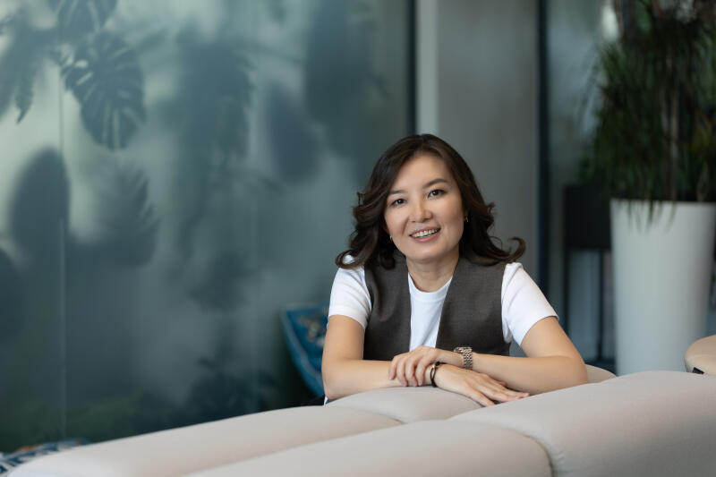 A smiling woman with dark hair, wearing a white t-shirt and grey vest, leans on a light grey sofa. Blurred plant silhouettes are visible behind a frosted glass partition in the background, with a potted plant to the right.