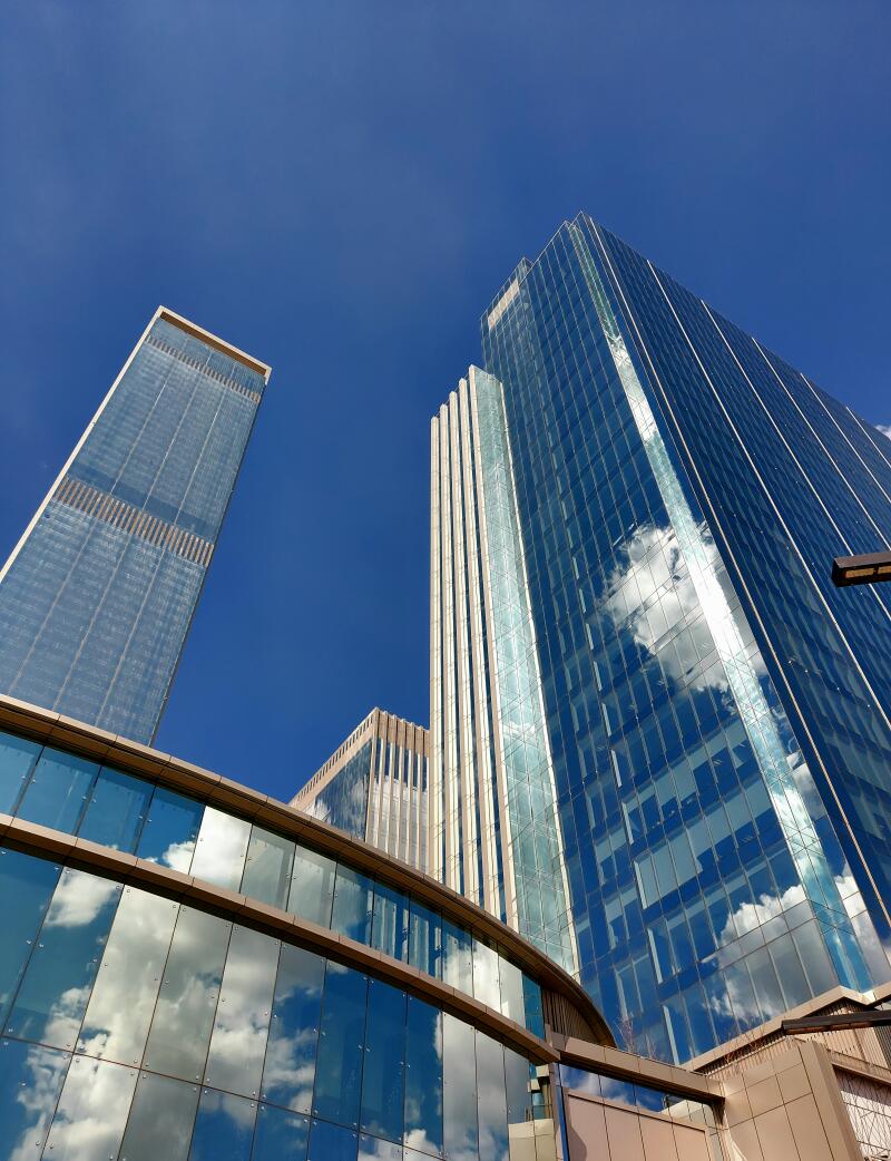 A low-angle shot of several modern skyscrapers with glass facades reflecting the blue sky and clouds.