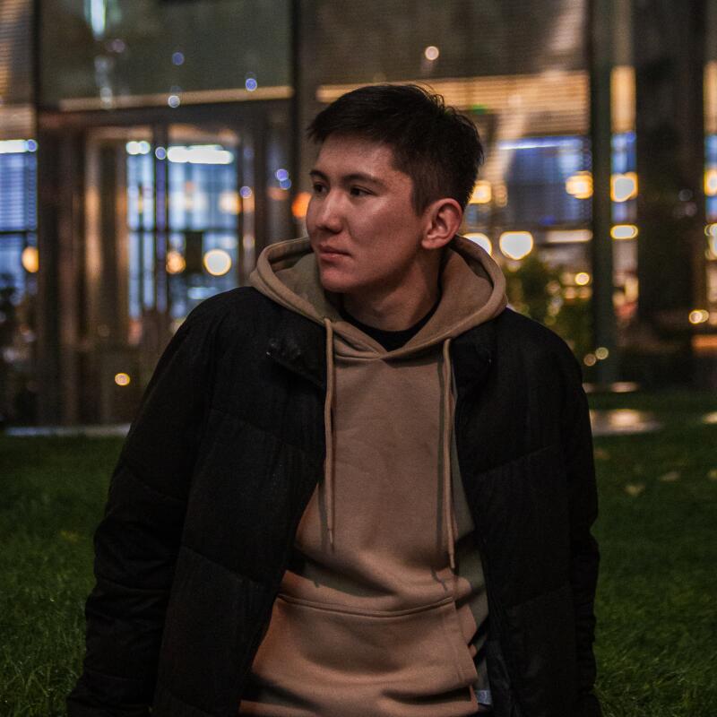 A young man in a black puffer jacket and beige hoodie looks to the side in an outdoor setting at night.