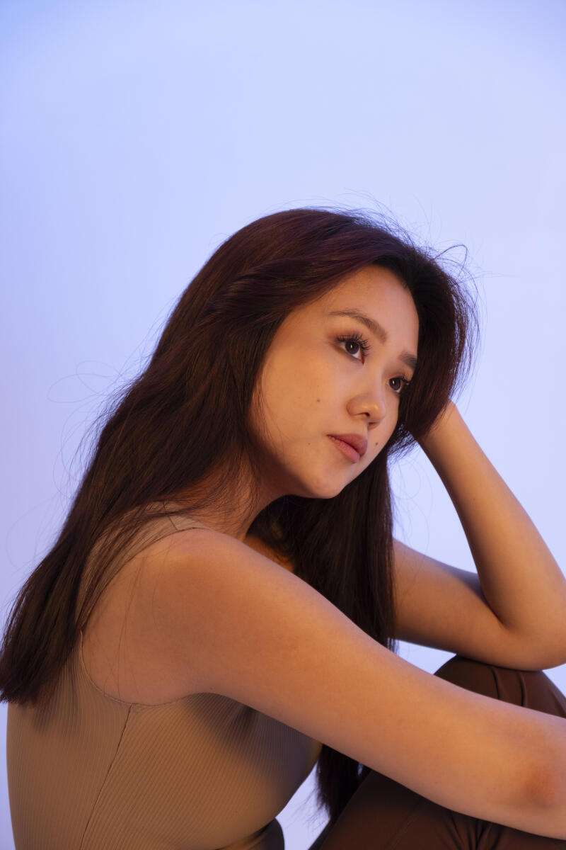 A woman with long, dark hair, wearing a tan tank top and brown pants, poses with her head resting on her hand against a light blue background.