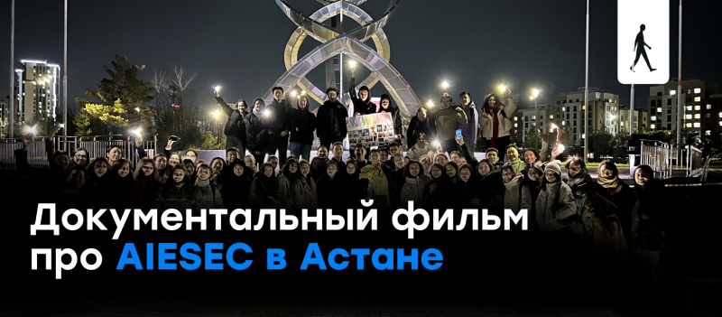 Группа людей стоит ночью перед современной художественной структурой, некоторые держат телефоны; текст гласит: «Документальный фильм про AIESEC в Астане».
