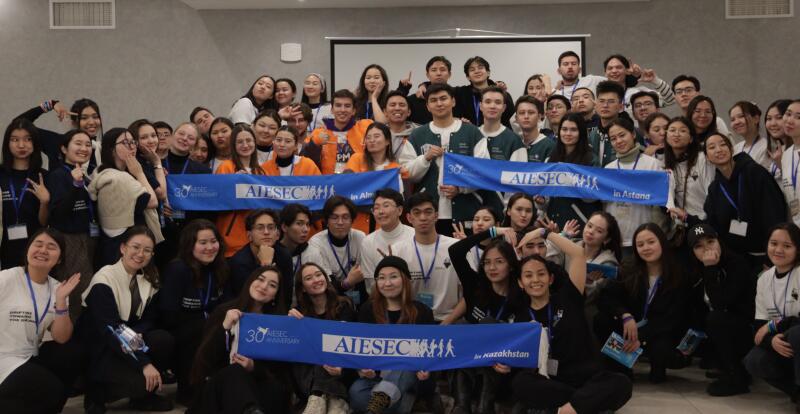 Большая группа молодых людей позирует для фотографии, держа баннеры с надписью «AIESEC» и «30 Anniversary». Группа разнообразна и, похоже, находится на мероприятии или конференции.