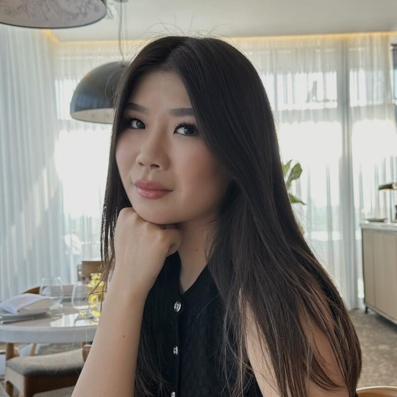 A woman with long, dark hair is sitting at a table, resting her chin on her hand. She is wearing a black sleeveless top with buttons.