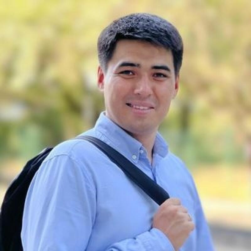 A man with dark hair and a light blue button-down shirt is holding a black backpack strap and smiling.