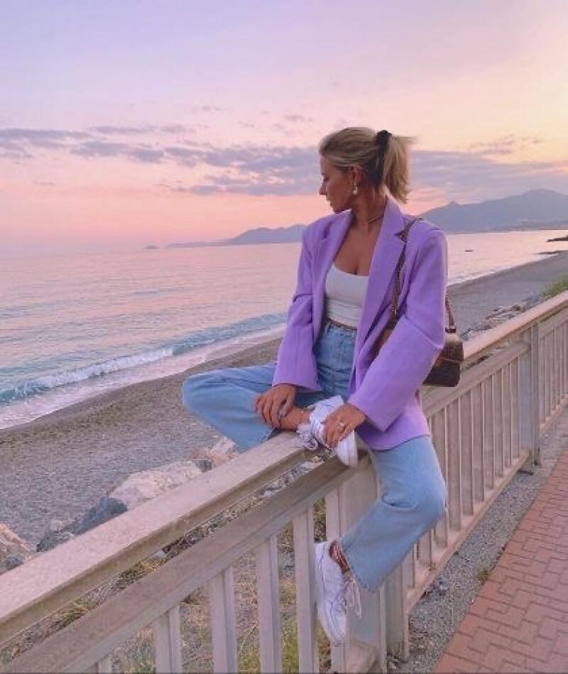 A woman wearing a lavender blazer and jeans sits on a railing overlooking the beach and ocean at sunset.