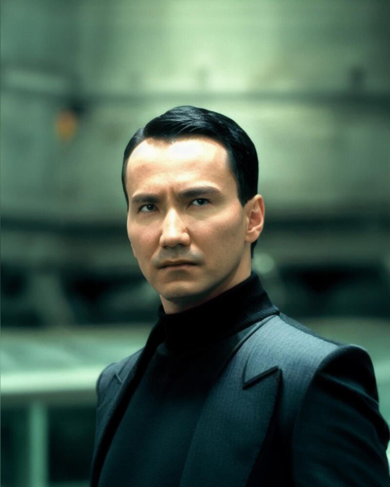 A man with dark hair and a serious expression is wearing a black turtleneck and a dark suit jacket.