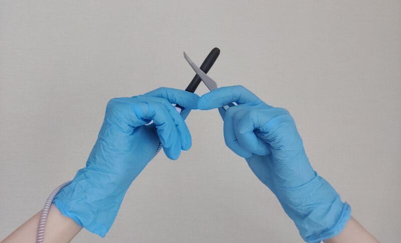 Two hands wearing blue surgical gloves hold a black and silver instrument in a crossed position against a light background.