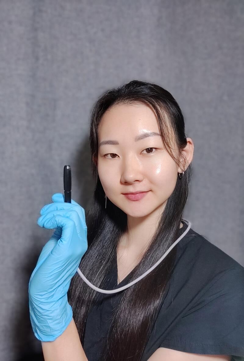 A woman in blue gloves holds a black pen, wearing a black shirt and a gray cord around her neck, against a gray background.