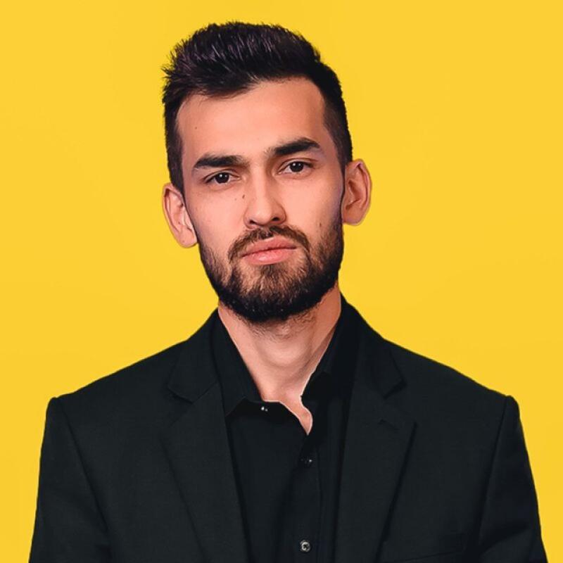 A man with dark hair and a beard is wearing a black suit against a yellow background.