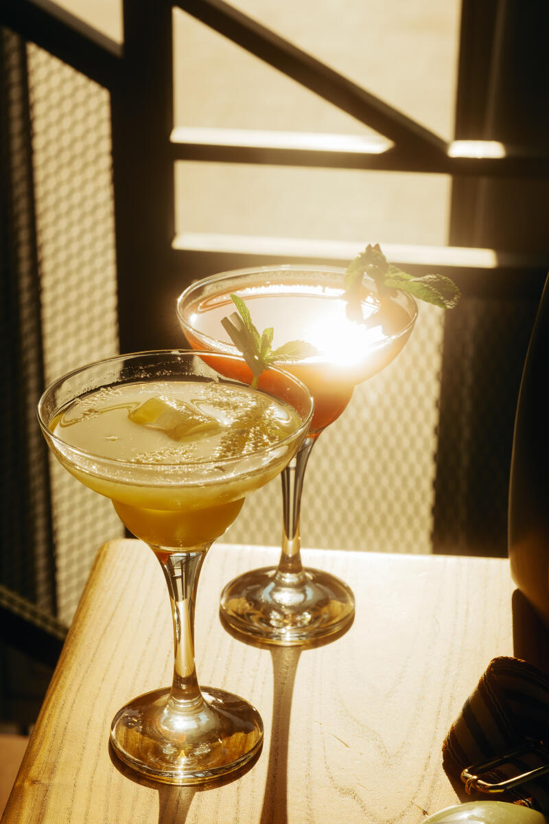 Two cocktails in martini glasses, one yellow and one red, are garnished with mint leaves and sit on a wooden surface.