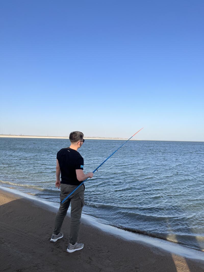 A man with a fishing rod stands on the beach looking at the water.