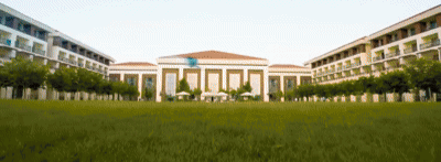 The image shows a large building with a tiled roof, framed by two multi-story buildings, all set against a backdrop of a green lawn and trees.