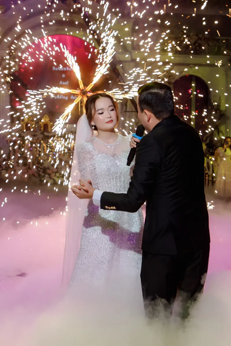 A bride in a white gown stands with a man in a black suit who is holding a microphone, amidst a backdrop of fireworks and fog.