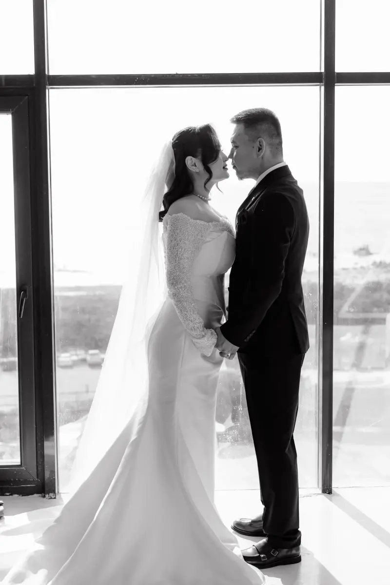 A black and white photo of a bride and groom standing in profile, holding hands and leaning in towards each other, against a bright window. The bride wears a white dress and veil, and the groom wears a dark suit.