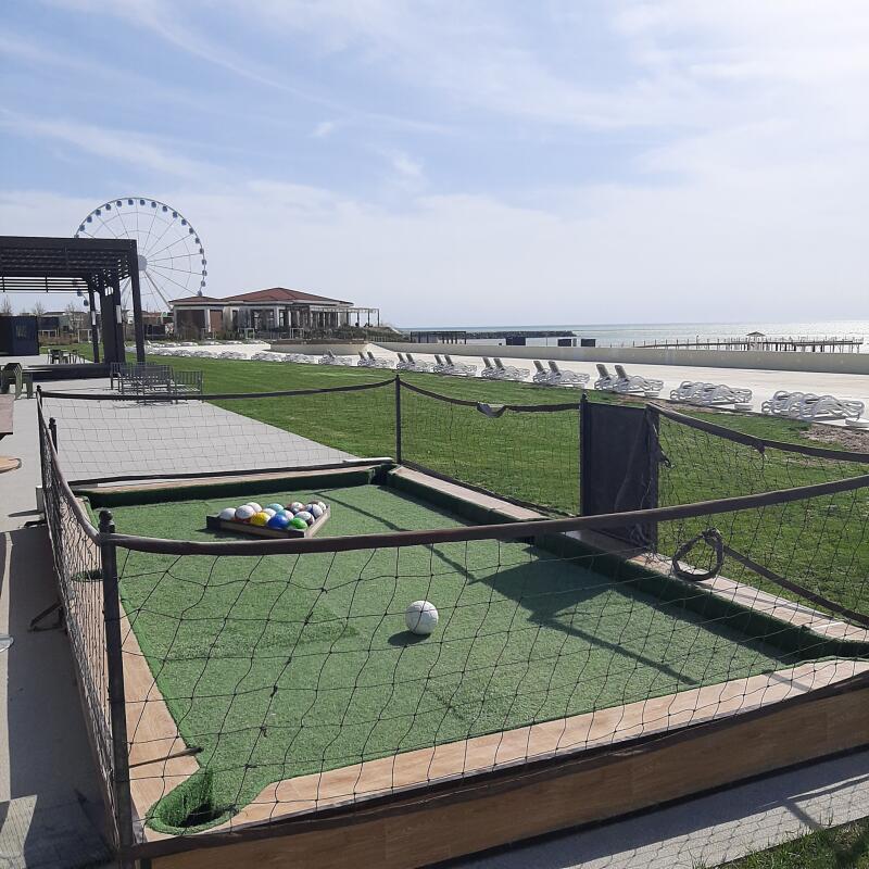 The image shows a snookball table with a set of balls racked in a triangle and a cue ball in the center, surrounded by a net fence and with a ferris wheel and beach in the background.